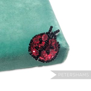 May include: A ladybug brooch made of red sequins and black beads, attached to a teal velvet surface. The brooch is a detailed, handcrafted accessory. The image includes the text "PETERSHAMS".