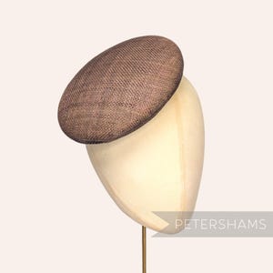 Op de afbeelding: Een bruine, geweven hoed met een ovale vorm, tentoongesteld op een mannequinhoofd. De hoed heeft een textuur en is waarschijnlijk gemaakt van natuurlijke vezels. Het mannequinhoofd is lichtbeige van kleur. De tekst "PETERSHAMS" is zichtbaar.