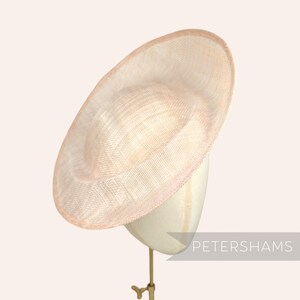 May include: A pale pink, wide-brimmed hat with a rounded crown and a delicate, woven texture. The hat is displayed on a mannequin head, showcasing its elegant design. The brim has a subtle pink trim.
