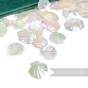 Peut inclure: Séquins irisés en forme de coquillage dispersés sur une surface blanche. Les sequins ont un éclat nacré, reflétant des couleurs comme le rose, le vert et le bleu. Un tissu vert est visible dans le coin supérieur gauche. Le texte "PETERSHAMS" est en bas à droite.