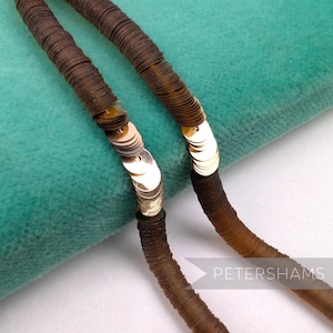 May include: Close-up of a brown sequin necklace with gold accents. The necklace is made of small, round sequins strung together. The background is a teal-colored fabric. The word "PETERSHAMS" is visible in the bottom right corner.