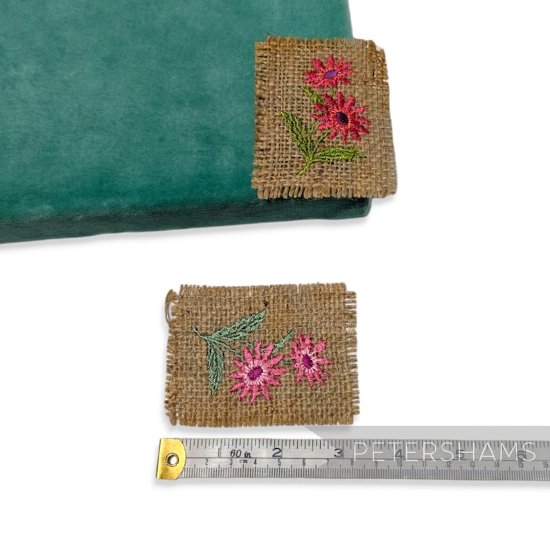 Pode incluir: Duas pequenas pe&ccedil;as quadradas de serapilheira com flores rosa bordadas e folhas verdes. Uma pe&ccedil;a est&aacute; presa a um tecido verde-azulado. Uma fita m&eacute;trica est&aacute; na imagem.