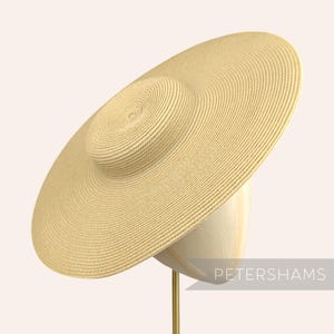 Może przedstawiać: Beżowy kapelusz przeciwsłoneczny z szerokim rondem i okrągłą koroną, wykonany z tkanego materiału. Kapelusz jest prezentowany na głowie manekina. Widoczny jest tekst "PETERSHAMS".