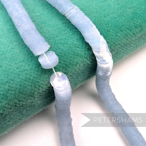 May include: Close-up of light blue sequin trim. The trim is made of small, round, iridescent sequins sewn together in a continuous line. The sequins are a pale blue color and have a slight sheen. The trim is shown against a green background.