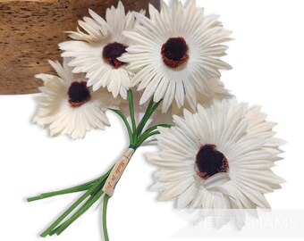 May include: A bunch of five white paper daisies with brown centers and green stems. The daisies are tied together with a small tag that reads "Made in Japan".