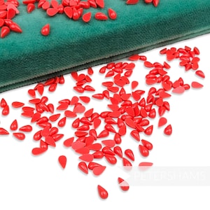 Può includere: Una collezione di perline di cera rosse a forma di goccia, sparse su una superficie bianca, con una sezione di tessuto di velluto verde sullo sfondo. Le perline sono lucide e uniformemente colorate.