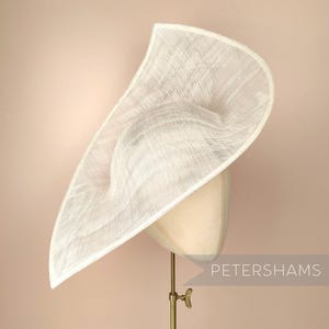 May include: A white, wide-brimmed hat made of woven straw, with a slightly pointed crown. The hat is displayed on a stand with the text "PETERSHAMS" visible in the lower right corner.