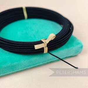 Peut inclure: Une bobine de cordon élastique noir, enroulée sur elle-même et attachée avec une étiquette en papier brun. Le cordon est posé sur une surface en velours bleu turquoise.
