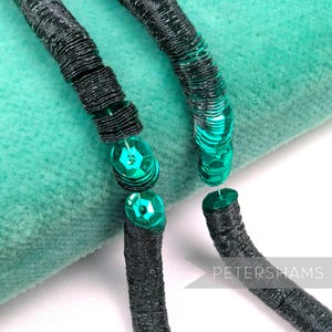 Peut inclure: Gros plan sur un collier avec des paillettes noires et turquoise. Le collier présente un design texturé et superposé avec des perles à facettes de couleur turquoise. Le fond est un tissu doux de couleur turquoise. Le mot "PETERSHAMS" est visible en bas à droite.