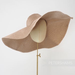 May include: A large, beige sun hat with a wide brim, displayed on a mannequin head. The hat has a woven texture and is supported by a gold-colored stand. The brim curves gracefully, offering ample shade. The word "PETERSHAMS" is visible in the lower right corner.