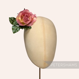 Può includere: Un manichino color crema adornato da una rosa rosa e gialla e foglie verdi. La rosa, con i suoi petali sfumati, è l'elemento principale. Il manichino è montato su un sottile supporto dorato. Il testo "PETERSHAMS" è visibile.