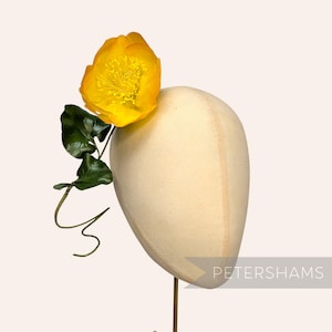 Puede incluir: Un maniquí de sombrerería color crema adornado con una flor amarilla brillante y hojas verdes. La flor está colocada en el lado izquierdo del maniquí. El texto "PETERSHAMS" es visible en la parte inferior derecha. Tocado elegante.