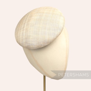 May include: A small, round, off-white hat made of woven straw. The hat is sitting on a white hat stand. The hat stand has a gold metal rod.