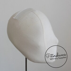May include: A white fabric millinery block with a clear plastic comb attached to the top. The block is on a black stand. Petershams Millinery Supplies is printed on the bottom right corner of the image.
