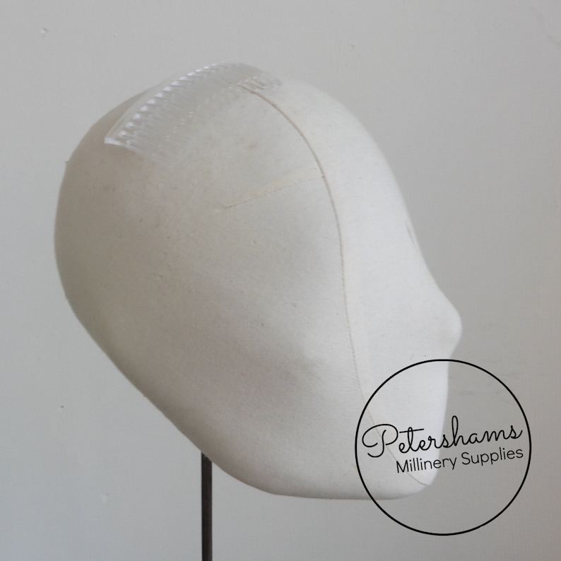May include: A white fabric millinery block with a clear plastic comb attached to the top. The block is on a black stand. Petershams Millinery Supplies is printed on the bottom right corner of the image.