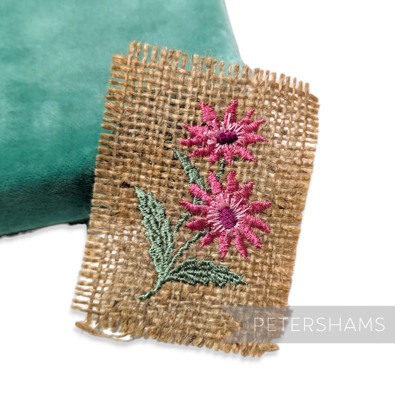 May include: A square of hessian fabric with an embroidered floral design. The embroidery shows two pink flowers with purple centres and green leaves. The hessian has a natural, woven texture.