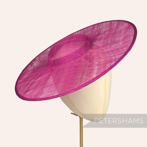 May include: A large, wide-brimmed hat made of pink straw. The hat is sitting on a white hat stand.