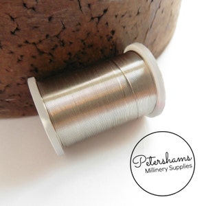 Könnte beinhalten: Eine Spule mit Silberdraht für Hutmacher, Basteln und Schmuckherstellung. Der Draht ist auf einer weißen Kunststoffspule aufgewickelt. Die Spule ist mit "Petershams Millinery Supplies" beschriftet.