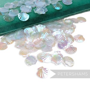 Pode incluir: Confetes iridescentes em forma de concha espalhados sobre uma superfície de veludo verde. Os confetes têm um brilho perolado, refletindo cores de rosa, azul e amarelo. A palavra "PETERSHAMS" está no canto inferior direito.