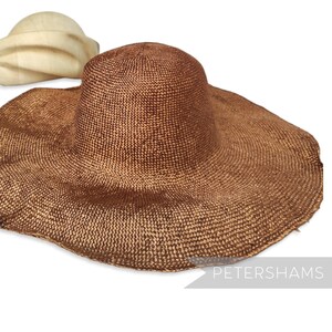 May include: A large, brown woven sun hat with a wide brim and a rounded crown. The hat is made of a textured material and has a natural, earthy appearance. A smaller, cream-coloured hat is visible in the background.