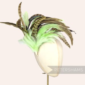 May include: A decorative headpiece featuring a mix of brown, green, and chartreuse feathers. The feathers are arranged on a cream-colored base, mounted on a stand. The text "PETERSHAMS" is visible.