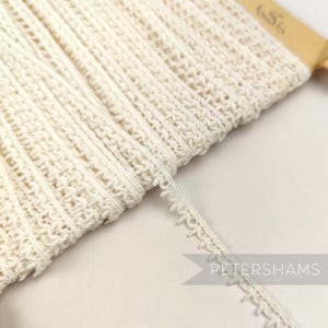 May include: A close-up of a white crocheted trim with a scalloped edge. The trim is folded and stacked on a wooden surface. The text "Petershams" is visible in the lower right corner of the image.