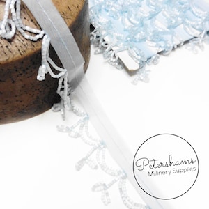 May include: A spool of light blue beaded fringe trim with a sheer backing. The trim is wrapped around a wooden spool. Petershams Millinery Supplies is printed on a white circle.