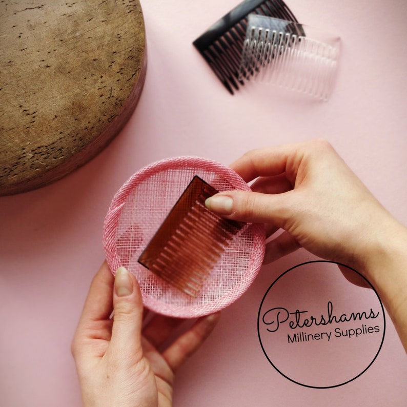 May include: A pink sinamay hat base with a brown comb being placed inside. The hat base is being held by a hand. Petershams Millinery Supplies is written in a circle in the bottom right corner of the image.