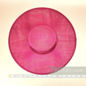 May include: A large, bright pink, round hat made of woven straw. The hat has a slightly indented centre and a smooth, even texture. A ruler is placed below the hat for scale.