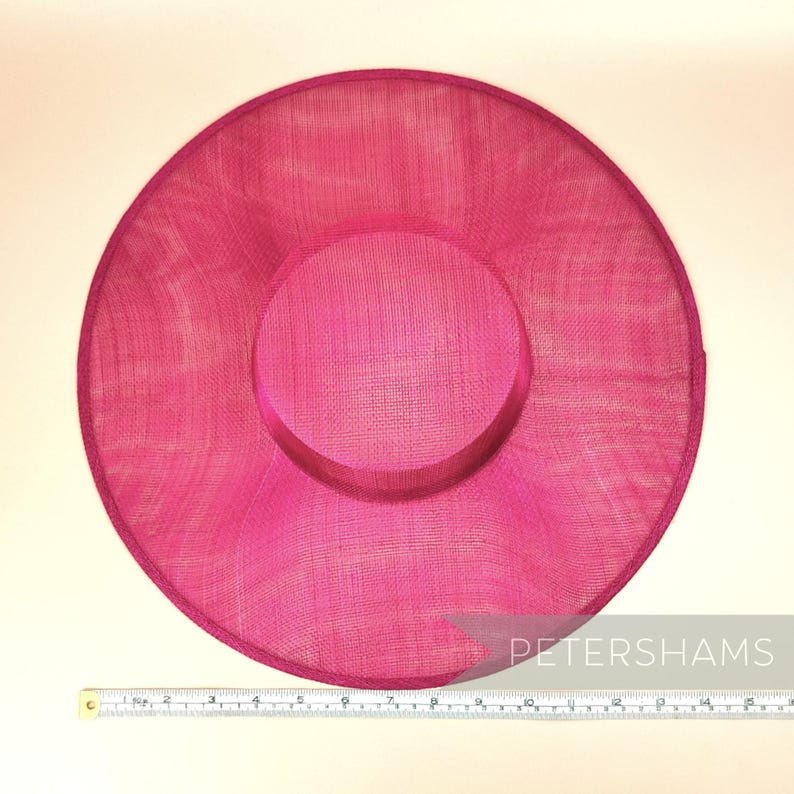 May include: A large, bright pink, round hat made of woven straw. The hat has a slightly indented centre and a smooth, even texture. A ruler is placed below the hat for scale.