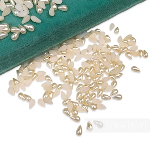 Può includere: Una grande quantità di perline a forma di lacrima madreperlacee color bianco avorio sono sparse su una superficie bianca. Le perline si riversano da una superficie di velluto verde.