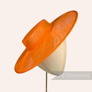 May include: A large, bright orange, wide-brimmed hat made of woven straw. The hat is sitting on a white hat form.