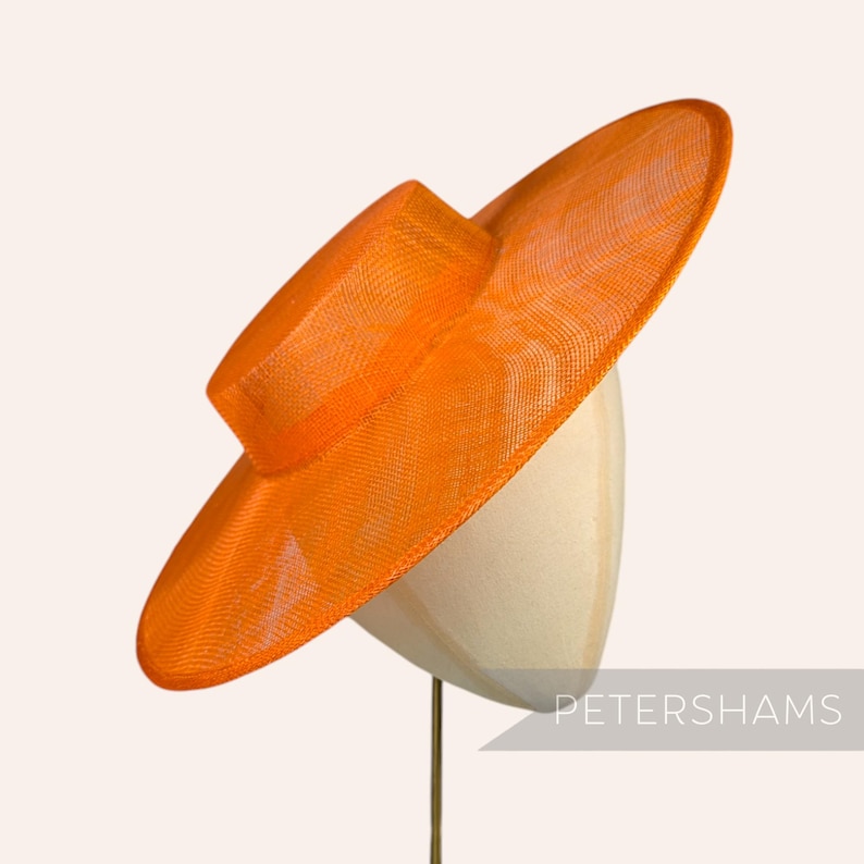 May include: A large, bright orange, wide-brimmed hat made of woven straw. The hat is sitting on a white hat form.