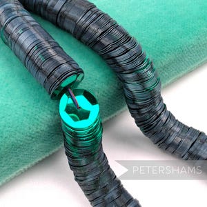 Op de afbeelding: Close-up van twee strengen donkerblauwe pailletten. De pailletten zijn cirkelvormig en aan elkaar geregen, met een heldere turquoise zeshoekige paillet aan het einde. De achtergrond is een zachte, lichtblauwe stof. Het woord "PETERSHAMS" staat in de rechter benedenhoek.