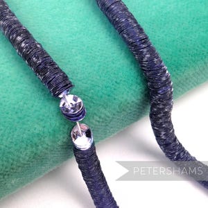 May include: Close-up of a dark blue beaded necklace with silver-colored connectors. The beads are flat and circular, with a slightly textured appearance. The necklace is displayed on a green velvet surface. The word "PETERSHAMS" is visible in the bottom right corner.