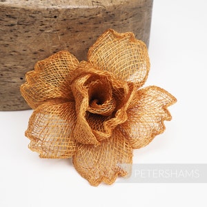 May include: A golden-bronze mesh flower, possibly a millinery accent, sits against a white background and a wooden block. The flower's petals are intricately woven, creating a textured, open design. The word "PETERSHAMS" is printed in white.