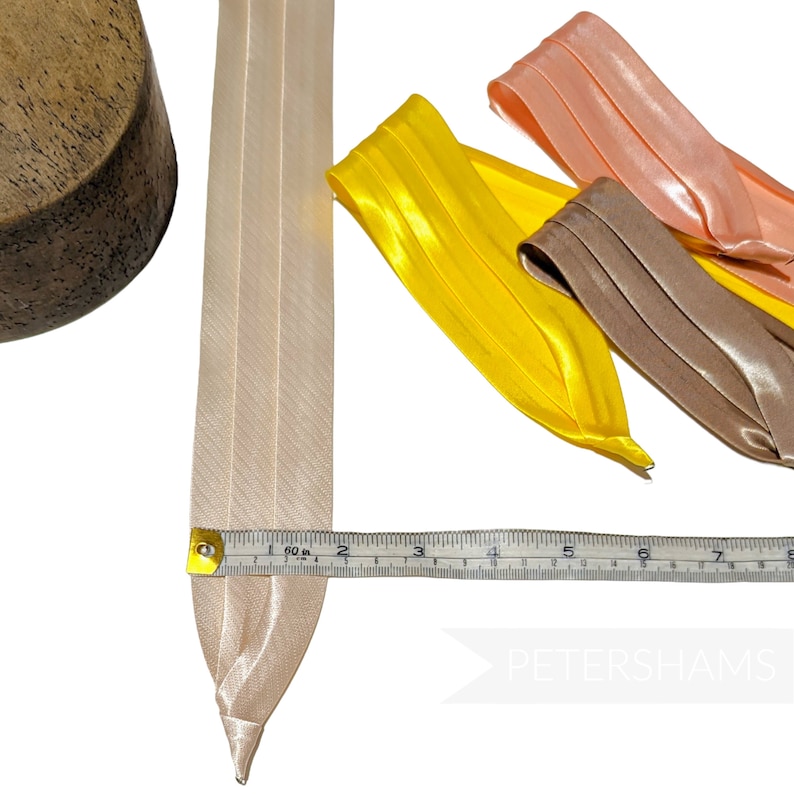 May include: A close-up of a beige satin ribbon measuring 7 inches long. The ribbon is lying on a white surface next to a ruler. Three other ribbons in yellow, peach, and brown are also visible in the image. The text "PETERSHAMS" is visible in the lower right corner of the image.