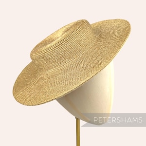 May include: A wide-brimmed, straw hat in a golden hue, displayed on a mannequin head. The hat features a structured, circular crown and a broad, downward-sloping brim. The hat is supported by a gold-colored stand. The text "PETERSHAMS" is visible.