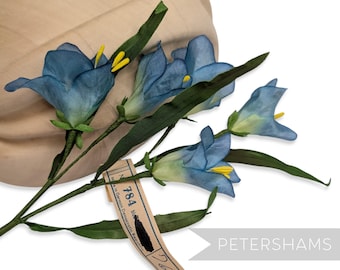 May include: Blue fabric flowers with yellow stamens and green stems are arranged near a beige hat. A tag with the number 784 and text in German is attached to the stems. The word "PETERSHAMS" is also visible.