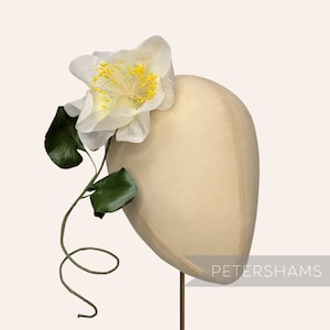 May include: A cream-coloured hat form decorated with a large, delicate white flower with yellow stamens and green leaves. The flower and leaves are attached to a curved stem. The word "PETERSHAMS" is printed in the lower right corner.