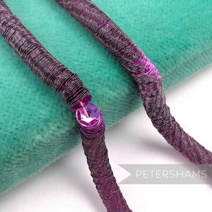 May include: Close-up of two strands of dark purple sequin trim. The trim is made of tightly wound, textured material with a line of sequins. A large, faceted sequin is attached to the end of one strand. The trim is on a teal green background.
