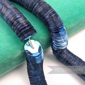 May include: Close-up of a blue sequin trim, featuring a series of connected, circular sequins. The trim is partially unraveled, revealing a wooden dowel. The background is a soft, green fabric. The text "PETERSHAMS" is visible in the lower right corner.