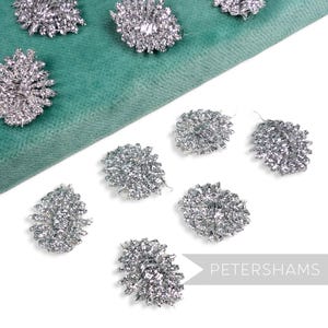 Peut inclure: Ornements métalliques argentés en forme de fleur, éparpillés sur une surface vert sarcelle. Les ornements sont faits de fil métallique et présentent un design délicat et complexe. Le mot "PETERSHAMS" est imprimé en blanc sur fond noir.