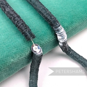 May include: Close-up of two strands of dark gray sequins, one partially unraveled, connected by a silver clasp. The sequins are tightly sewn together, creating a textured, cylindrical shape. The background is a soft, teal-colored fabric.