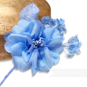 May include: A light blue fabric flower with multiple petals and a cluster of white beads at the center. The flower is accompanied by smaller blue blossoms and a pale blue leaf. The stem is a matching light blue color.