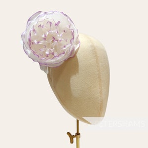 May include: A white and purple fabric flower with ruffled edges, displayed on a beige hat form.
