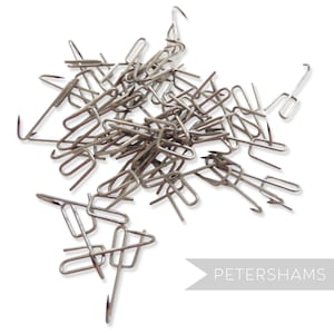 May include: A pile of silver metal hooks, commonly used for attaching fabric to a garment. The hooks are in a jumbled heap, with some of the hooks pointing upwards and others pointing downwards.