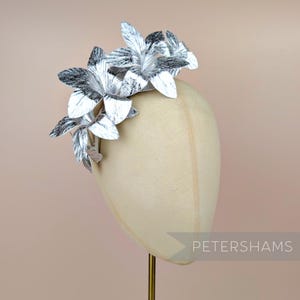 May include: A silver floral headpiece with multiple flower accents. The headpiece is displayed on a mannequin head. The flowers have a textured appearance and are in shades of silver and gray. The word "PETERSHAMS" is visible.
