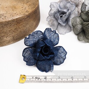 May include: Close-up of navy blue, gray, and olive green fabric flower embellishments. A wooden form is in the background. A measuring tape shows the flowers are approximately 4 inches in diameter. The flowers are likely for millinery or craft projects.