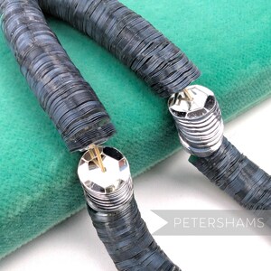 May include: A close-up of a dark gray necklace with a unique design. The necklace is made of small, round, metallic pieces strung together. The clasp is silver and geometric. The word "PETERSHAMS" is in the bottom right corner.